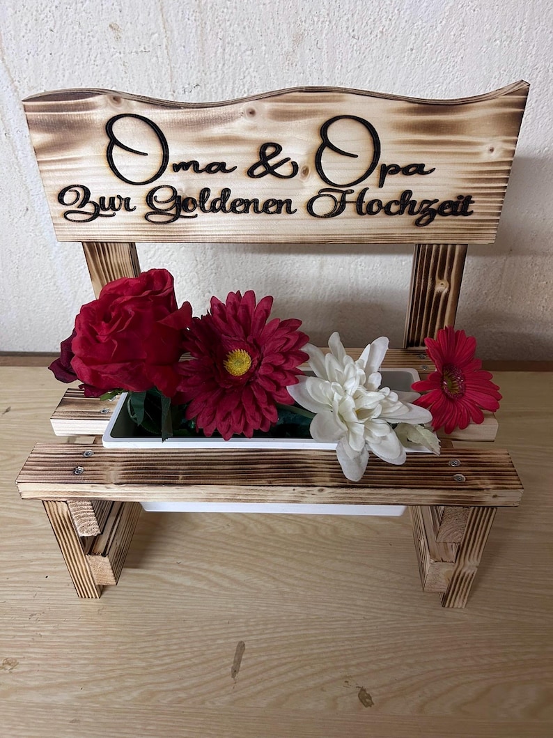K&ouml;nnte beinhalten: Ein holzbankf&ouml;rmiger Pflanzk&uuml;bel mit den Worten "Oma & Opa Zur Goldenen Hochzeit" in schwarzer Schrift. Der Pflanzk&uuml;bel enth&auml;lt rote, wei&szlig;e und rosa Blumen. Das Holz hat eine gebrannte Oberfl&auml;che.