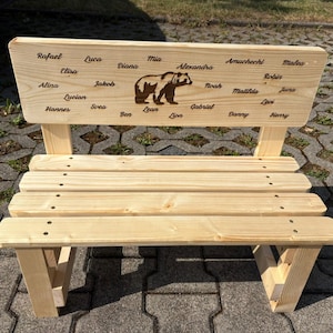 May include: A small, light-colored wooden bench with a backrest featuring a bear illustration and engraved names. The bench has a slatted seat and angled legs. The names include Rafael, Luca, Mia, and others.