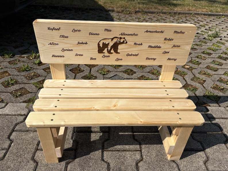 May include: A small, light-colored wooden bench with a backrest featuring a bear illustration and engraved names. The bench has a slatted seat and angled legs. The names include Rafael, Luca, Mia, and others.