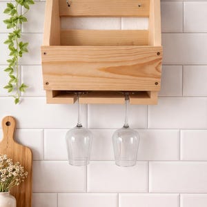 Puede incluir: Un botellero de madera natural con dos copas de vino colgando debajo. El estante est&aacute; montado en una pared de azulejos blancos. Una tabla de cortar de madera y un jarr&oacute;n con flores est&aacute;n en la encimera.