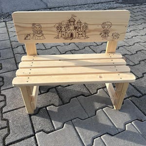 May include: A small, light-colored wooden bench with a backrest featuring engraved illustrations of a princess, a castle, and a prince. The bench has a slatted seat and angled legs, suitable for children.