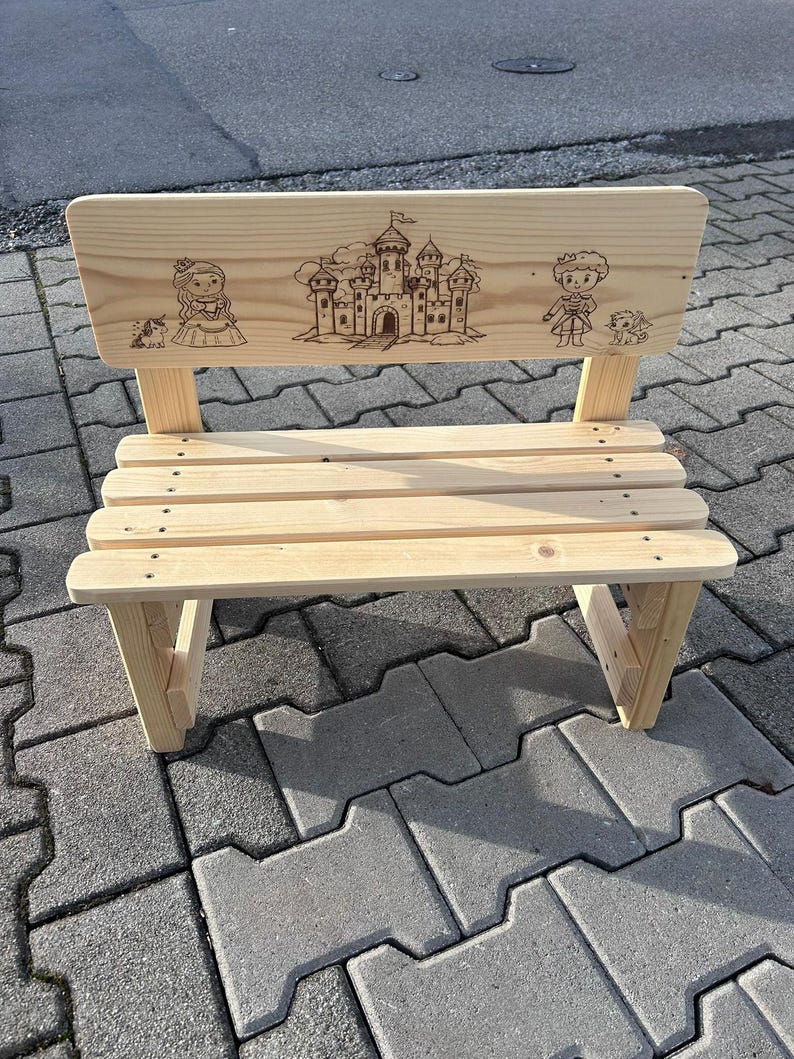 May include: A small, light-colored wooden bench with a backrest featuring engraved illustrations of a princess, a castle, and a prince. The bench has a slatted seat and angled legs, suitable for children.