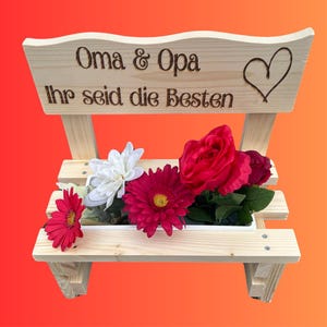 K&ouml;nnte beinhalten: Ein holzbankf&ouml;rmiger Pflanzk&uuml;bel mit den Worten "Oma & Opa Ihr seid die Besten" und einem Herzen auf der R&uuml;ckseite. Der Pflanzk&uuml;bel enth&auml;lt rote, wei&szlig;e und rosa Blumen.
