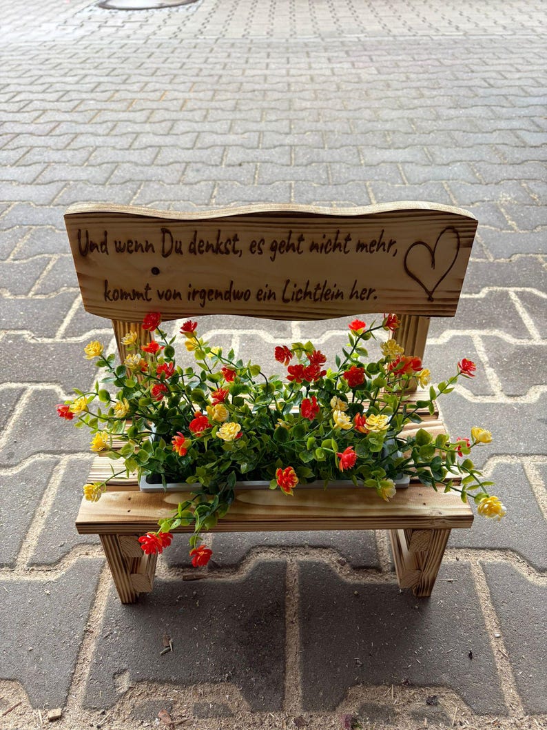 K&ouml;nnte beinhalten: Ein kleiner Holzbank-Blumentopf mit R&uuml;ckenlehne. Die R&uuml;ckenlehne tr&auml;gt den Text "Und wenn Du denkst, es geht nicht mehr, kommt von irgendwo ein Lichtlein her." und ein Herz. Die Bank enth&auml;lt einen Pflanztopf mit roten und gelben Blumen.