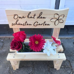 K&ouml;nnte beinhalten: Ein holzbankf&ouml;rmiger Pflanzk&uuml;bel mit dem deutschen Text "Oma hat den sch&ouml;nsten Garten" und einem Blumendesign. Der Pflanzk&uuml;bel enth&auml;lt rote Rosen, eine rosa Gerbera, eine wei&szlig;e Blume und eine rote Blume.