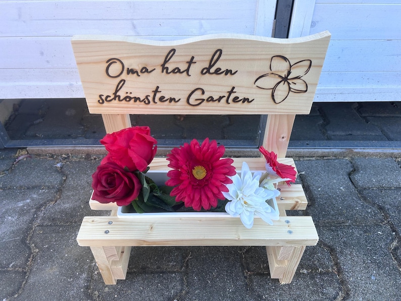 K&ouml;nnte beinhalten: Ein holzbankf&ouml;rmiger Pflanzk&uuml;bel mit dem deutschen Text "Oma hat den sch&ouml;nsten Garten" und einem Blumendesign. Der Pflanzk&uuml;bel enth&auml;lt rote Rosen, eine rosa Gerbera, eine wei&szlig;e Blume und eine rote Blume.