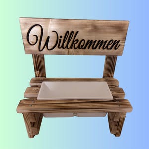 K&ouml;nnte beinhalten: Eine rustikale Holzbank mit dem Wort "Willkommen" in schwarzer Schrift. Die Bank hat eine rechteckige wei&szlig;e Schale und ist aus hellbraunem Holz mit sichtbarer Maserung gefertigt.