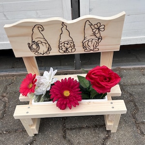 K&ouml;nnte beinhalten: Ein Blumenkasten im Stil einer Holzbank mit drei Zwergenillustrationen auf der R&uuml;ckseite. Der Pflanzkasten enth&auml;lt rote, wei&szlig;e und magentafarbene Blumen. Die Bank ist aus unbehandeltem Holz gefertigt und steht auf einer Ziegeloberfl&auml;che.