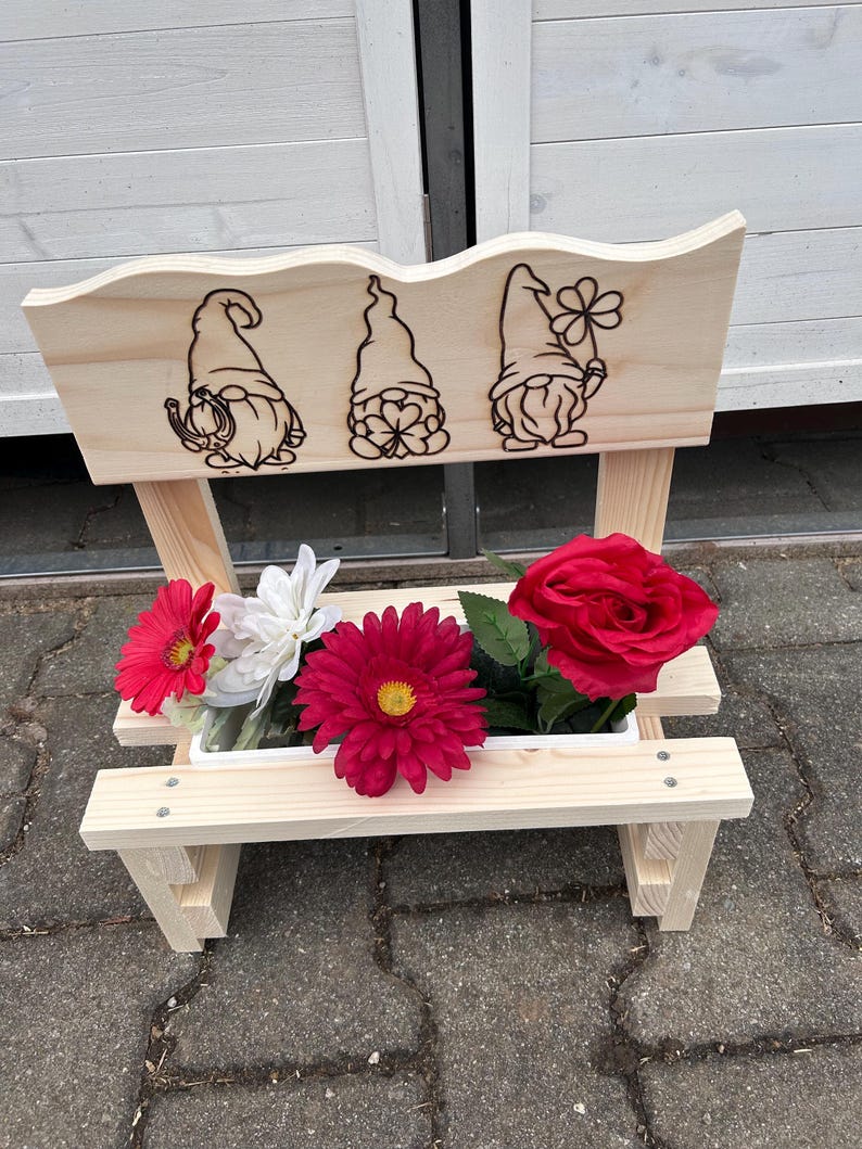 K&ouml;nnte beinhalten: Ein Blumenkasten im Stil einer Holzbank mit drei Zwergenillustrationen auf der R&uuml;ckseite. Der Pflanzkasten enth&auml;lt rote, wei&szlig;e und magentafarbene Blumen. Die Bank ist aus unbehandeltem Holz gefertigt und steht auf einer Ziegeloberfl&auml;che.