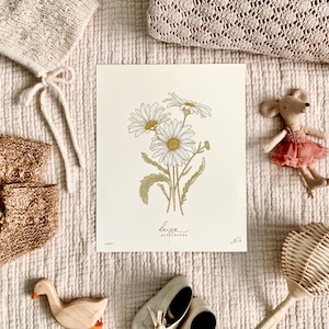 May include: A white and gold botanical print with a drawing of daisies and the text "Leuca Asteraceae". The print is on a white background and is surrounded by knitted baby items, a wooden toy duck, and a wooden rattle.