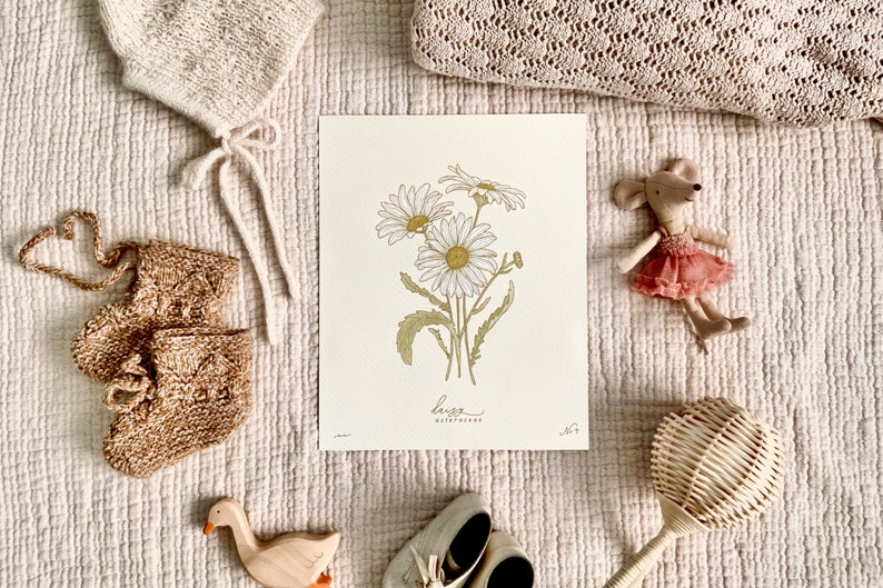 May include: A white and gold botanical print with a drawing of daisies and the text "Leuca Asteraceae". The print is on a white background and is surrounded by knitted baby items, a wooden toy duck, and a wooden rattle.
