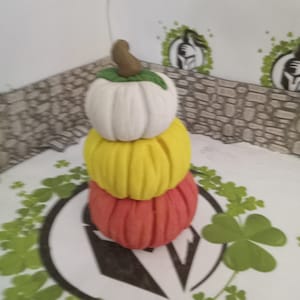 Può includere: Una pila di tre zucche in miniatura, la superiore bianca, la intermedia gialla e la inferiore arancione.