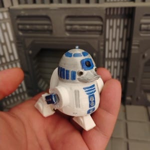 Könnte beinhalten: Eine kleine, 3D-gedruckte Figur eines weißen und blauen Droiden mit einem entenartigen Schnabel. Die Figur hat einen kuppelförmigen Kopf mit blauen und silbernen Akzenten, einen zylindrischen Körper mit blauen Details und kleine weiße Beine.