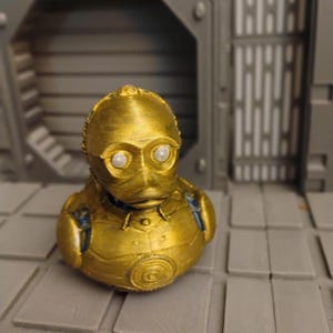 Könnte beinhalten: Eine goldfarbene, 3D-gedruckte Gummiente, die dem Charakter C-3PO nachempfunden ist. Die Figur hat ein metallisch goldenes Finish, einen abgerundeten Kopf mit zwei weißen, kreisförmigen Augen und eine detaillierte Brustplatte mit einem runden Emblem.