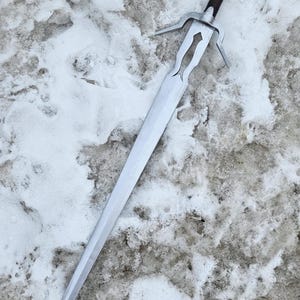 May include: A long, silver sword with a brown handle and a decorative crossguard. The sword is lying on a snowy surface, with the blade pointing towards the bottom of the frame. The handle has a circular pommel.