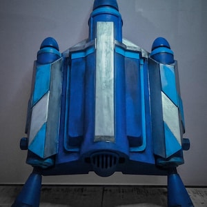 May include: A blue and silver prop jetpack with a central fin, two side boosters, and two lower thrusters. The jetpack has a detailed design with geometric shapes and metallic accents. The color scheme includes shades of blue and silver.