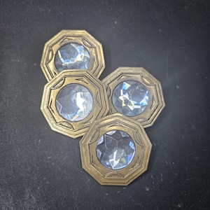 May include: Four gold-coloured, octagonal-shaped buttons with clear glass centres. The buttons have a decorative design around the edges.