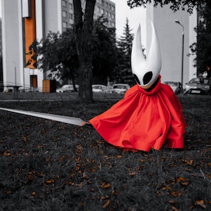 May include: A person in a white mask with black eye holes and a red cloak, holding a long silver sword. The person is sitting on the grass in front of a building.