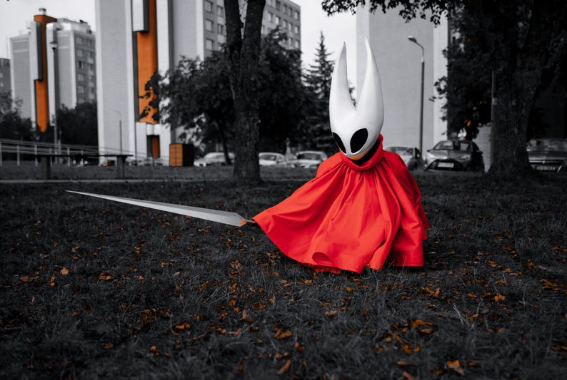 May include: A person in a white mask with black eye holes and a red cloak, holding a long silver sword. The person is sitting on the grass in front of a building.