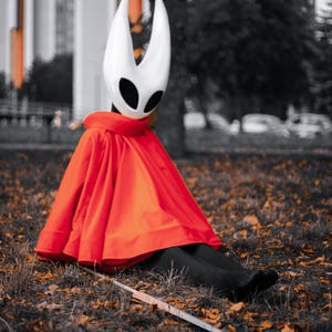 May include: A person wearing a white mask with black eye holes and a red cape, sitting on the ground with a sword. The mask has two upward-curving horns. The cape is a bright red color. The background is a blurred cityscape.