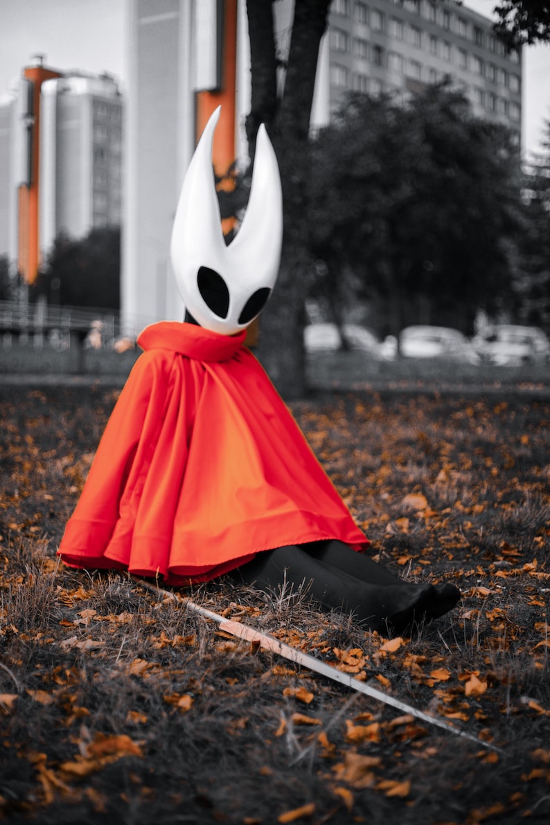 May include: A person wearing a white mask with black eye holes and a red cape, sitting on the ground with a sword. The mask has two upward-curving horns. The cape is a bright red color. The background is a blurred cityscape.