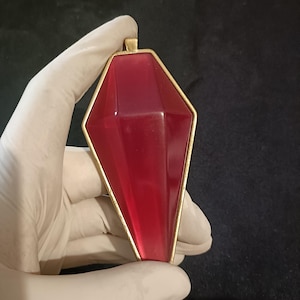 Puede incluir: Un colgante facetado de color rojo rubí con un marco dorado. El colgante tiene una forma geométrica alargada con un lazo en la parte superior para una cadena. El fondo es de color oscuro y liso.