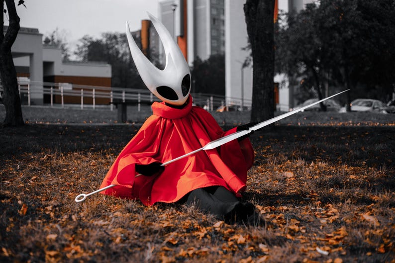 May include: A person in a cosplay costume of a character from the video game Hollow Knight. The costume includes a white mask with horns, a red cape, and a long, silver spear. The person is sitting on the ground covered in fallen leaves.