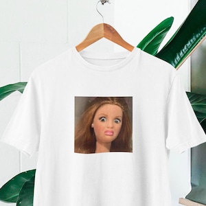 Könnte beinhalten: Weißes T-Shirt mit einem quadratischen Bild eines Puppengesichts mit überraschtem Ausdruck. Die Puppe hat rotes Haar und trägt rosa Lippenstift. Das T-Shirt hängt an einem Holzkleiderbügel.