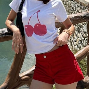 May include: A white t-shirt with a red cherry graphic print. The model is wearing red denim shorts.
