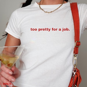 May include: White t-shirt with the text "too pretty for a job." in red lettering. The shirt is being worn by a person with a red crossbody bag and a gold necklace.