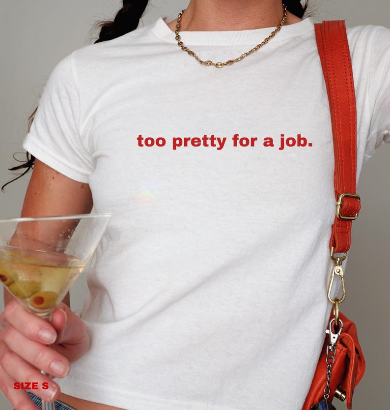 May include: White t-shirt with the text "too pretty for a job." in red lettering. The shirt is being worn by a person with a red crossbody bag and a gold necklace.