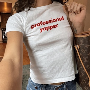 May include: White t-shirt with the red text "professional yapper". The short-sleeved shirt is paired with blue jeans. A brown handbag with a logo is visible in the background.