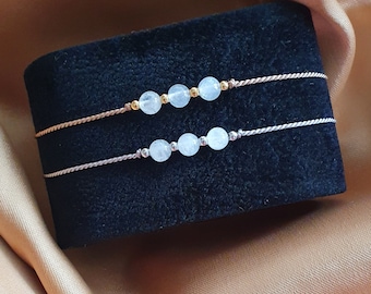 May include: Two delicate bracelets with light blue gemstone beads and thin brown cords. The top bracelet has gold beads between the gemstones.