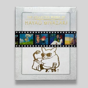 May include: A boxed set titled "The Collected Works of Hayao Miyazaki" with gold lettering. The set features a film strip design with animated scenes and a gold illustration of a pig wearing sunglasses. The box is white with a textured border.