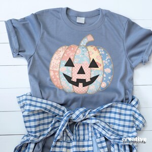 Pode incluir: Uma camiseta azul clara com um design de abóbora de retalhos com padrões florais rosa, azul e amarelo. A abóbora tem um rosto e um caule pretos. Uma camisa xadrez azul e branca está amarrada na cintura.