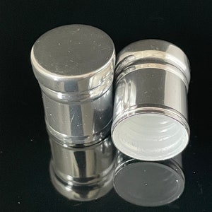 Puede incluir: Dos tapones de botella plateados con un acabado brillante. Los tapones son de forma cilíndrica con una parte superior ligeramente redondeada.