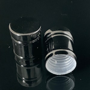Puede incluir: Dos tapones de botella negros con un acabado brillante. Un tapón está en posición vertical y el otro está tumbado de lado, revelando un interior de plástico blanco. Los tapones son cilíndricos y parecen estar hechos de metal o material similar.