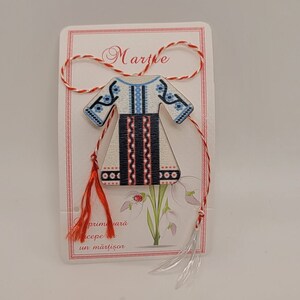 Pode incluir: Um amuleto martisor de madeira branca em forma de vestido tradicional romeno com bordados azuis e vermelhos. O amuleto est&aacute; preso a um cord&atilde;o vermelho e branco com borlas. O cart&atilde;o diz "Martie" e "primavara cepe un martisor".