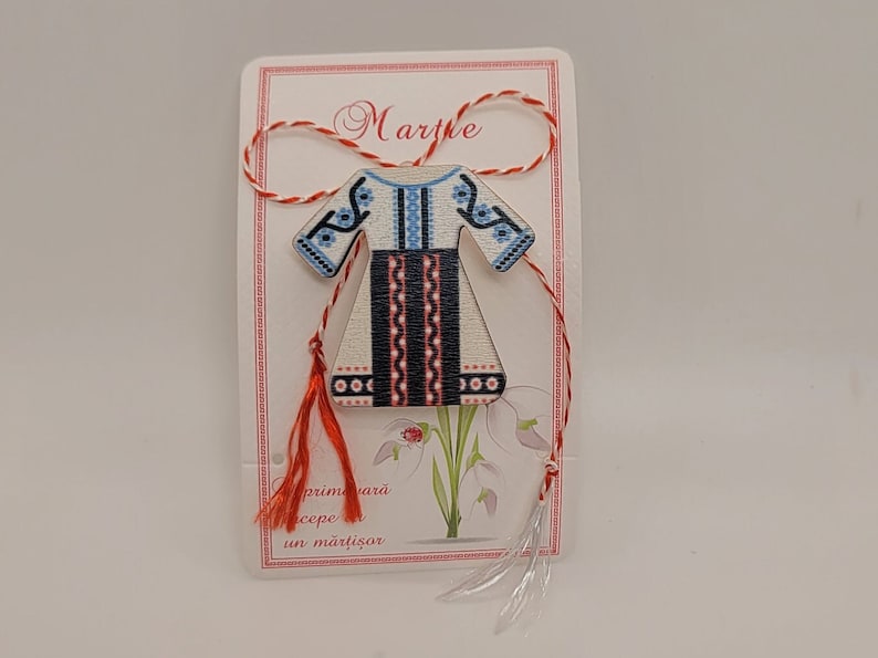 Pode incluir: Um amuleto martisor de madeira branca em forma de vestido tradicional romeno com bordados azuis e vermelhos. O amuleto est&aacute; preso a um cord&atilde;o vermelho e branco com borlas. O cart&atilde;o diz "Martie" e "primavara cepe un martisor".