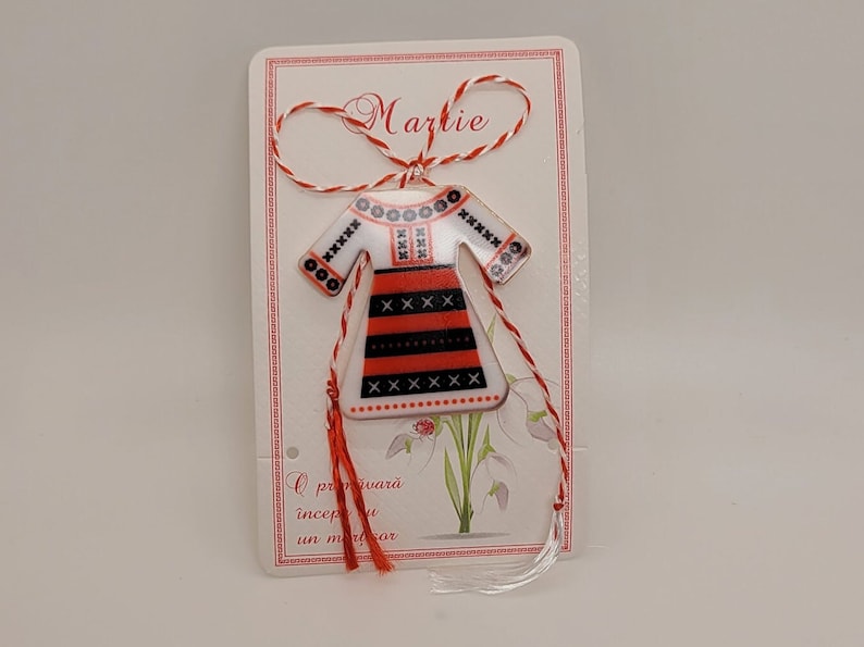 Pode incluir: Um cord&atilde;o vermelho e branco com um pequeno ornamento de cer&acirc;mica em forma de vestido tradicional romeno. O ornamento &eacute; decorado com bordados pretos e vermelhos. O cord&atilde;o &eacute; amarrado com um la&ccedil;o e o ornamento est&aacute; pendurado no cord&atilde;o. O cart&atilde;o diz "Martie" e tem uma mensagem em romeno.