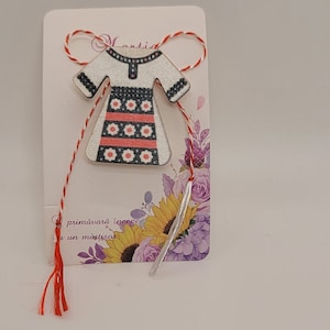 Pode incluir: Um ornamento de madeira branco em forma de vestido tradicional romeno com listras vermelhas e pretas e flores brancas. O ornamento &eacute; amarrado com um cord&atilde;o vermelho e branco com borlas. O cart&atilde;o diz "primavara incepe cu un martisor".