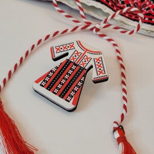 Pode incluir: Um broche de madeira branca em forma de vestido tradicional romeno com bordados vermelhos e pretos. O broche est&aacute; preso a um cord&atilde;o vermelho e branco com borlas.