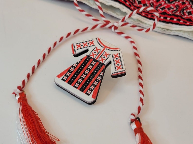 Pode incluir: Um broche de madeira branca em forma de vestido tradicional romeno com bordados vermelhos e pretos. O broche est&aacute; preso a um cord&atilde;o vermelho e branco com borlas.