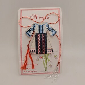 Pode incluir: Um amuleto martisor de madeira branca em forma de vestido tradicional romeno com bordados azuis e vermelhos. O amuleto est&aacute; preso a um cord&atilde;o vermelho e branco com borlas. O cart&atilde;o diz "Martie" e "primavara incepe cu un martisor".