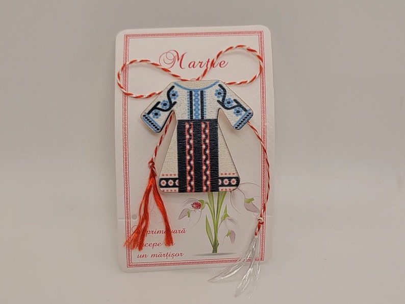 Pode incluir: Um amuleto martisor de madeira branca em forma de vestido tradicional romeno com bordados azuis e vermelhos. O amuleto est&aacute; preso a um cord&atilde;o vermelho e branco com borlas. O cart&atilde;o diz "Martie" e "primavara incepe cu un martisor".