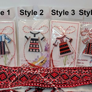 Pode incluir: Quatro estilos diferentes de amuletos romenos Martie, cada um com um recorte de madeira de um vestido tradicional romeno com diferentes padr&otilde;es de bordado. Os amuletos est&atilde;o presos a um cord&atilde;o vermelho e branco.
