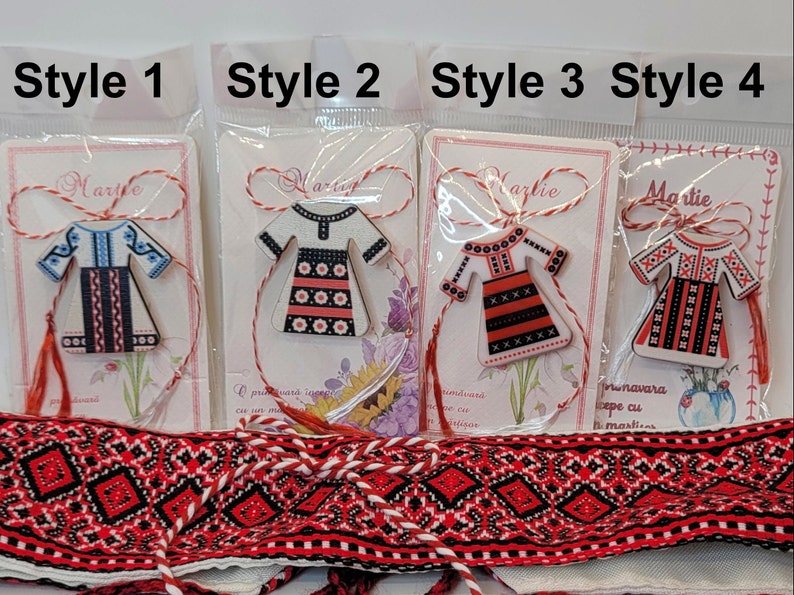 Pode incluir: Quatro estilos diferentes de amuletos romenos Martie, cada um com um recorte de madeira de um vestido tradicional romeno com diferentes padr&otilde;es de bordado. Os amuletos est&atilde;o presos a um cord&atilde;o vermelho e branco.