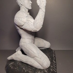 May include: A white painted figurine of a man kneeling on a rock base. The man's right arm is raised and his hand is open. The figurine is detailed with textured lines throughout.
