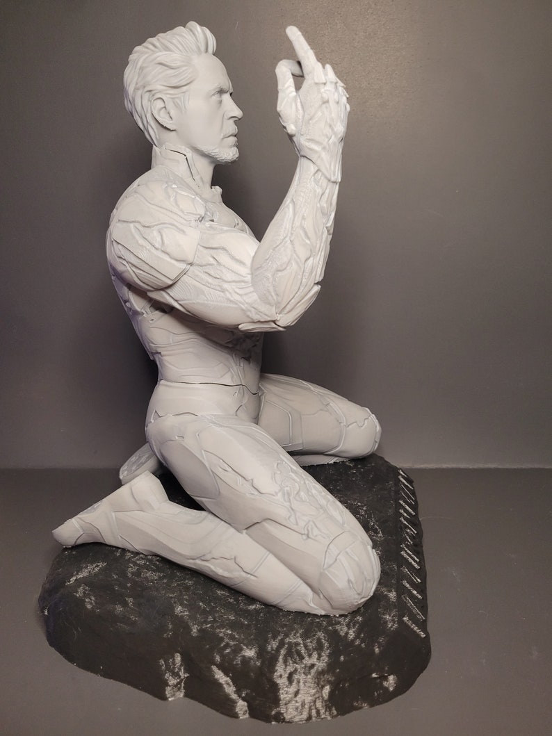 May include: A white painted figurine of a man kneeling on a rock base. The man's right arm is raised and his hand is open. The figurine is detailed with textured lines throughout.