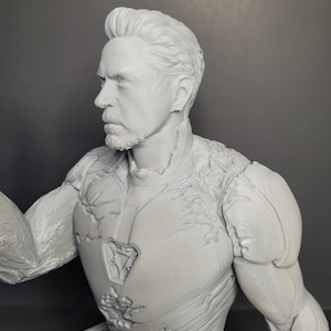 May include: A gray 3D printed figurine of a superhero wearing a detailed, textured suit. The figure has a determined expression and a beard.
